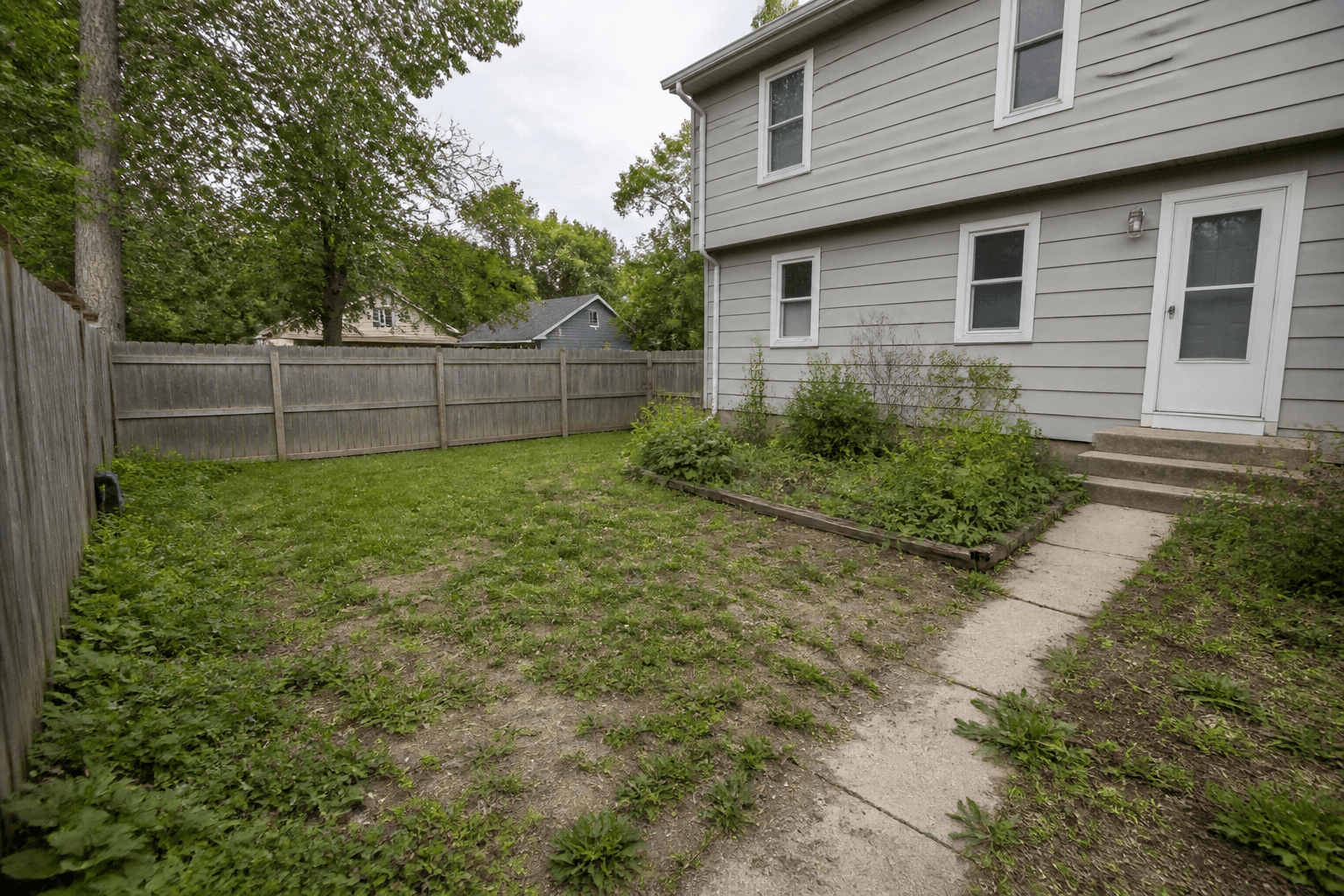 The same Kansas City backyard before renovation, with patchy grass, weeds, and a cracked concrete walkway